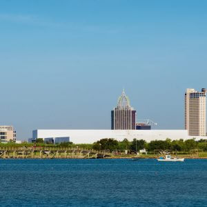 The skyline of Mobile, Alabama, with a short, wide building on the left, then 3 taller buildings to the right, with the river in the foreground.