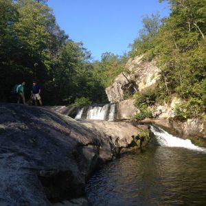 Wilson Creek in North Carolina, flowing over two small ridges with trees on either side and a blue sky above. Two people stand on the left side, one wearing a bright teal shirt, the other wearing a dark shirt and khaki shorts.