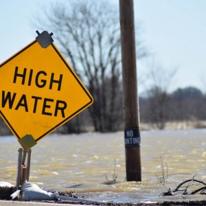 A yellow sign says "high water" in front of a flooded roadway backed by leafless trees.